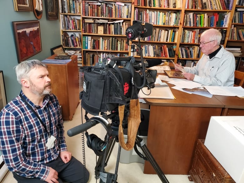 Two men sit in an office near a camera and a bookshelf. 