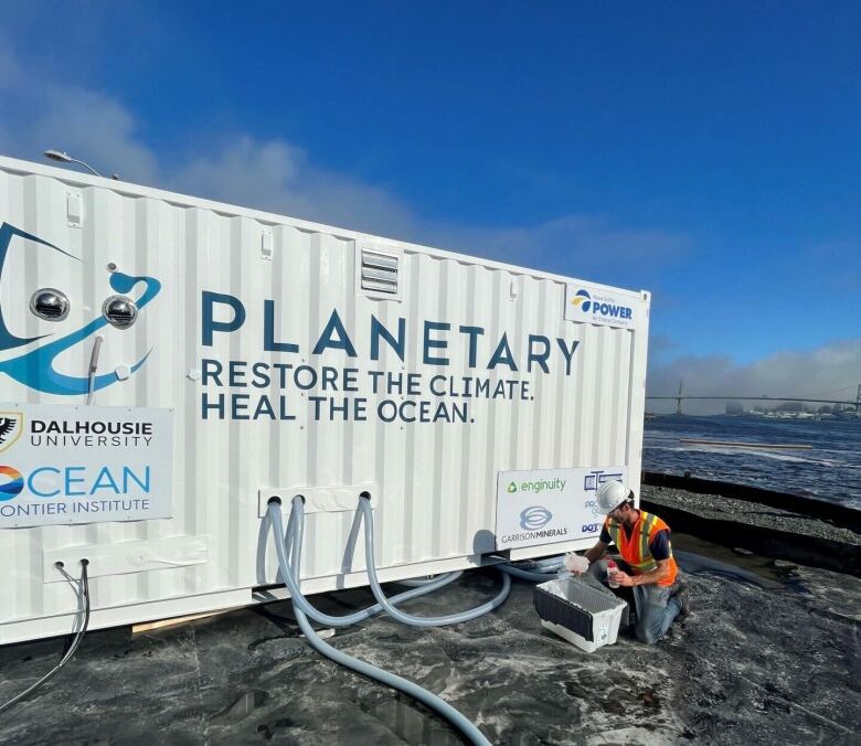 A man in a hard hat fills bottles in front of a shipping container with hoses protruding from it.