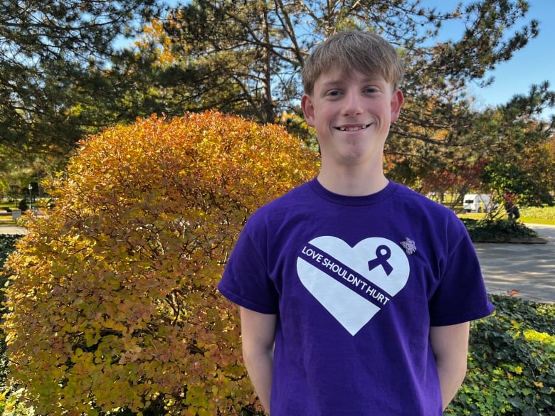 A boy smiles at the camera wearing a purple shirt that says 