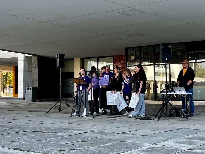 A group stands around a microphone wearing purple shirts and holding posters. 