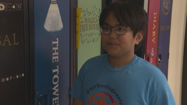 A young boy in a blue shirt with glasses stands in front of lockers.