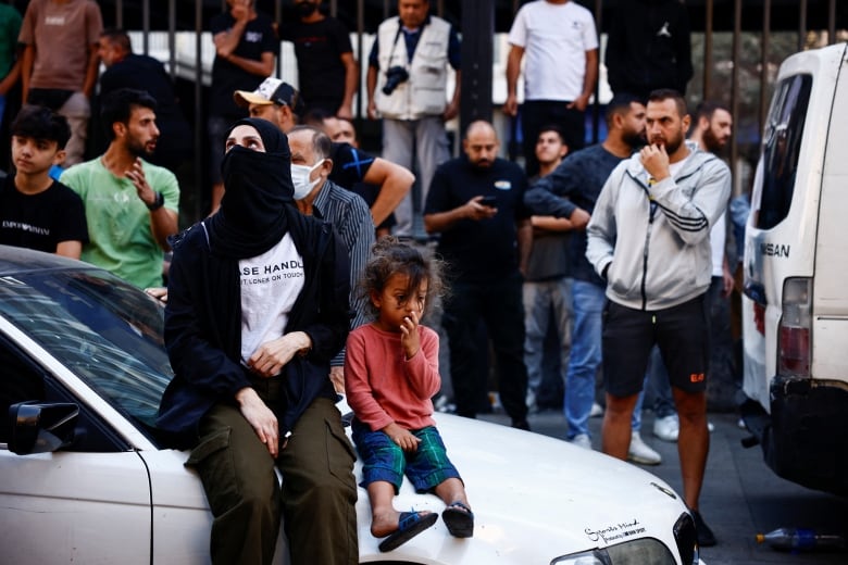 A woman and a child sit on the hood of a car among a crowd of people.