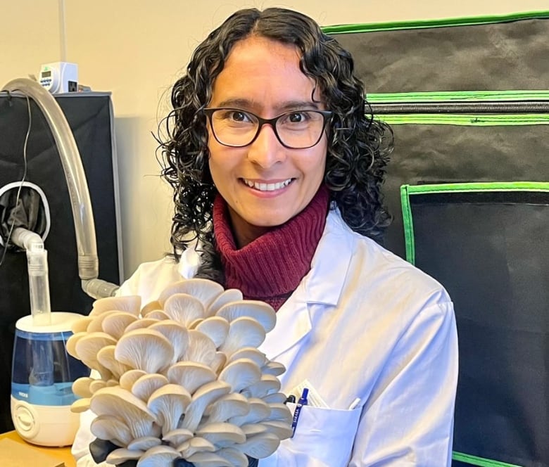 A woman wears a white lab coat and holds a bunch of mushrooms. 