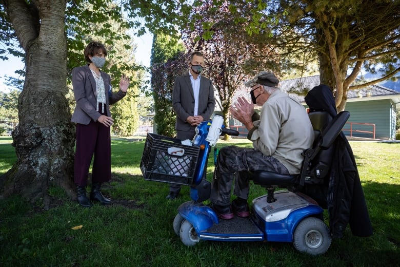 Two standing people in facemasks speak to a man seated on an electric scooter outdoors.