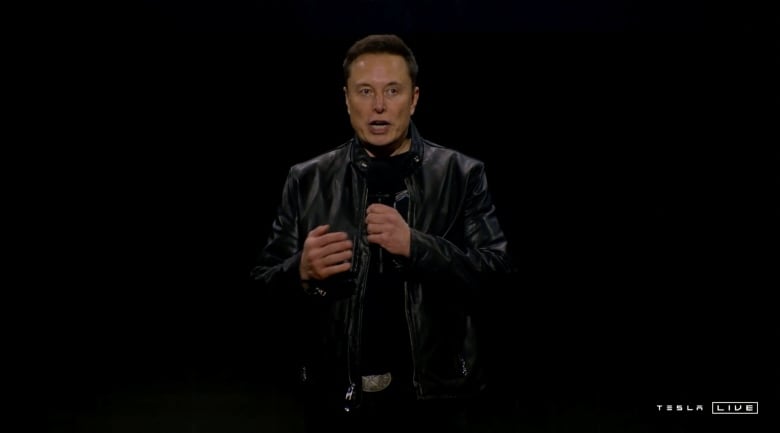 Elon Musk is pictured in a leather jacket speaking at an event. 