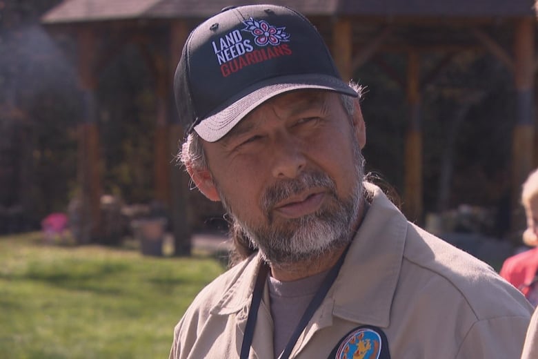 A man in a brown work shirt and ball cap that says 'land needs guardians' listens to someone speak.