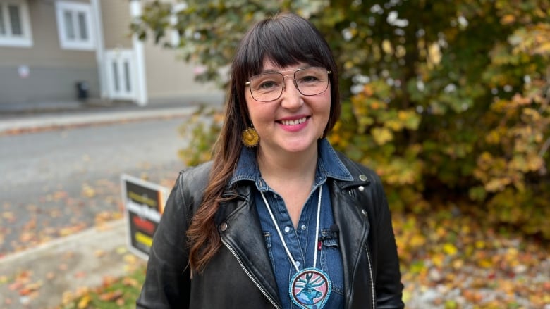 A smiling woman wearing glasses and a black leather jacket stands in front of a tree.