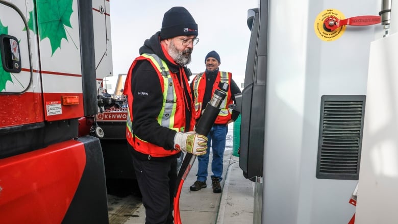 a man in red and black holds a gas hose as another man watches