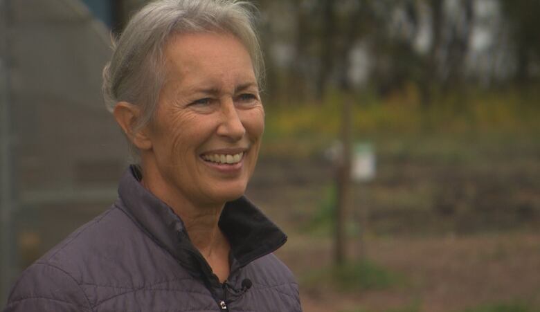 A woman smiles, a garden and greenhouse behind her.