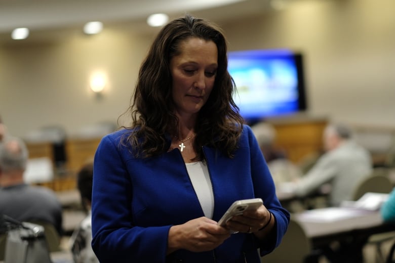 A woman in a business blazer and with a serious expression on her face looks down at a cellphone in her hands.