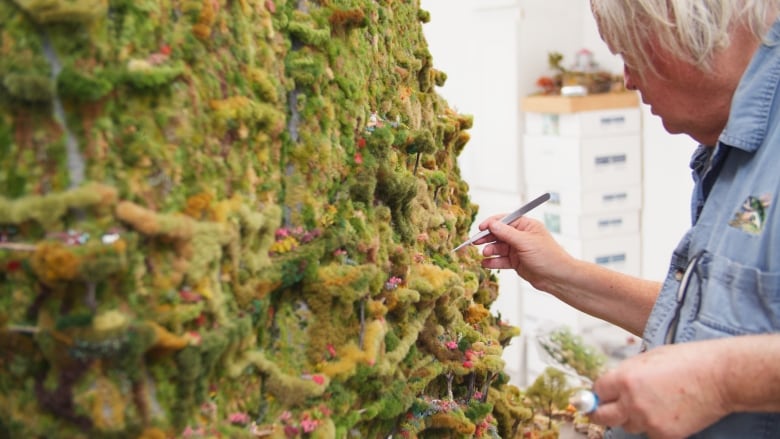 A man with long grey hair uses tweezers to make adjustments to a sculpture that resembles a gigantic mountain covered in greenery.