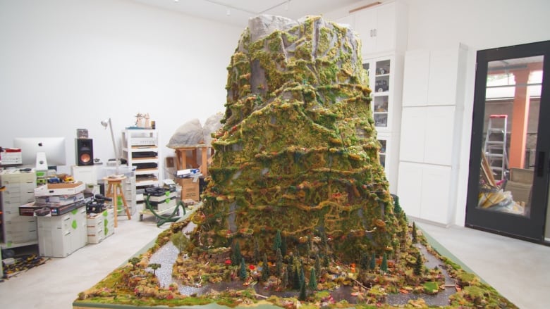 A lush 8-foot-tall mountain detailed with railroad modelling materials sits inside an artist's studio. 