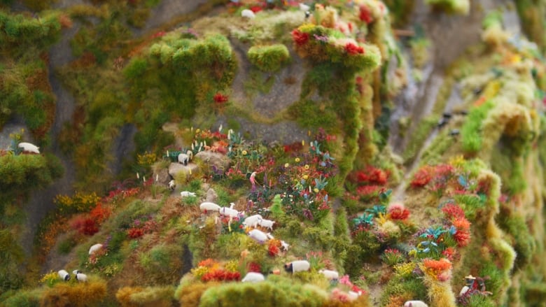 Built from railroad modelling materials, the scene shows a woman gardening flowers and other plants on a rugged landscape, surrounded by animals.