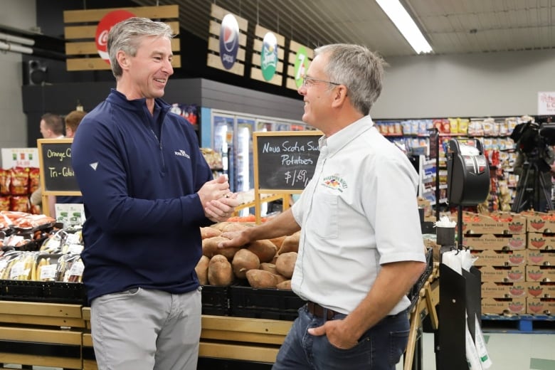 The premier holds his hands together in excitement while looking at another man who has one hand casually in his pocket.