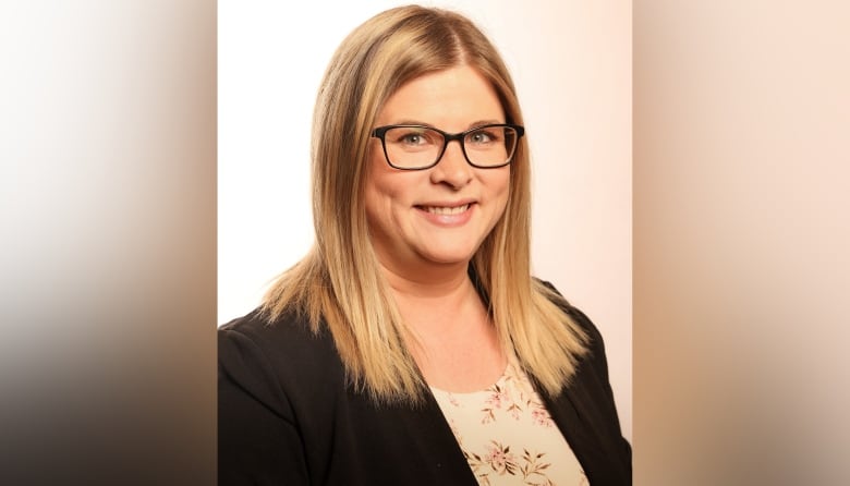 A woman with shoulder-length, blond hair wearing black-rimmed glasses and a black blazer smiles to the camera.