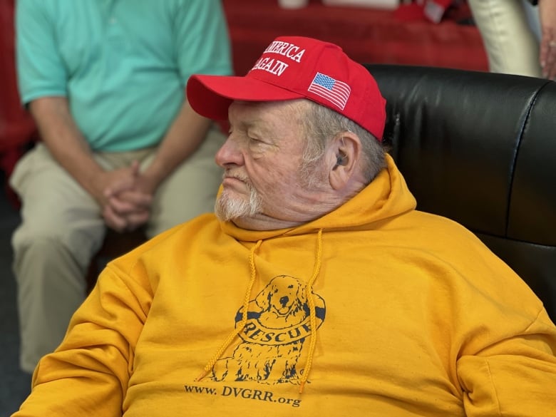 Older man with red MAGA cap, seated in chair 