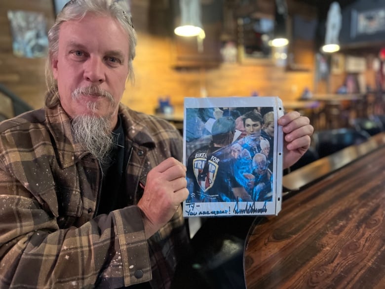 Man in bar holds up picture of a younger version of himself confronting a protester in a Bernie Sanders T-shirt, and the photo is signed in marker by Donald Trump with the message, 