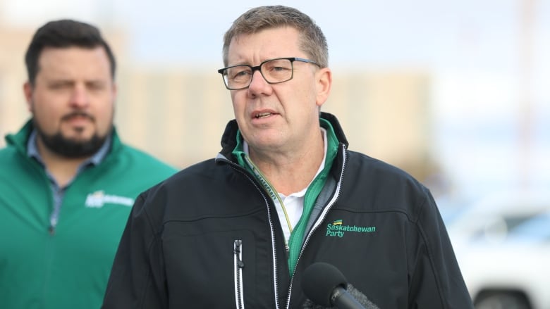 A man stands at a podium. The man is wearing a white shirt with a black jacket over top. The jacket has the phrase 