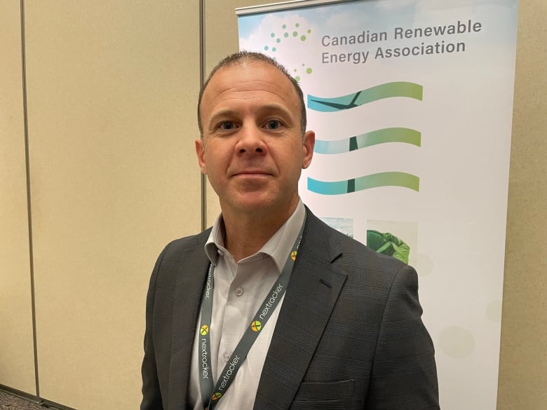 A man in a gray suit jacket and white button down shirt stands in front of a banner for the Canadian Renewable Energy Association.