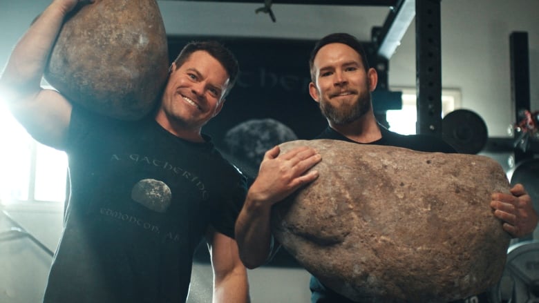 Two men hold very large stones.