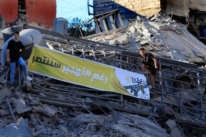 Two men stand on rubble holding a banner with Arabic printing that reads : 'Despite the displacement we will be victorious.'