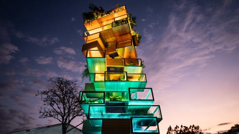 A tall, Jenga-shaped structure made of hollow boxes that are brightly lit in green, gold and blue, pictured from below against a purple dusk sky. Plant life spills out from each box.