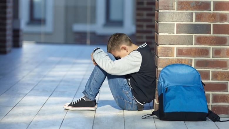 A young boy sits against a brick wall, his dead down on his folded knees, a blue backpack beside him. 