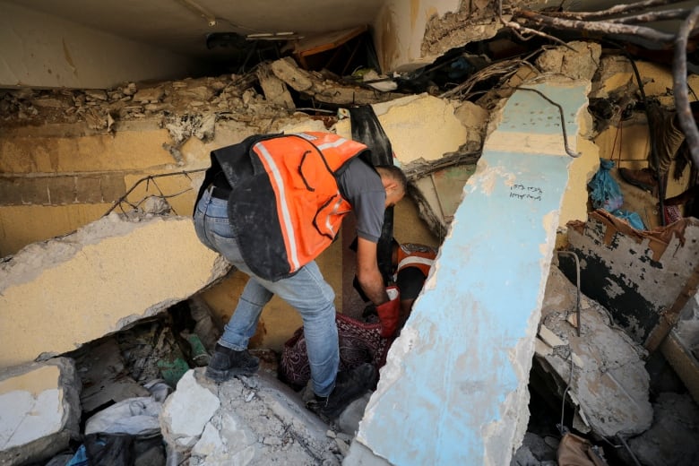 A man in an orange vest looks through rubble 