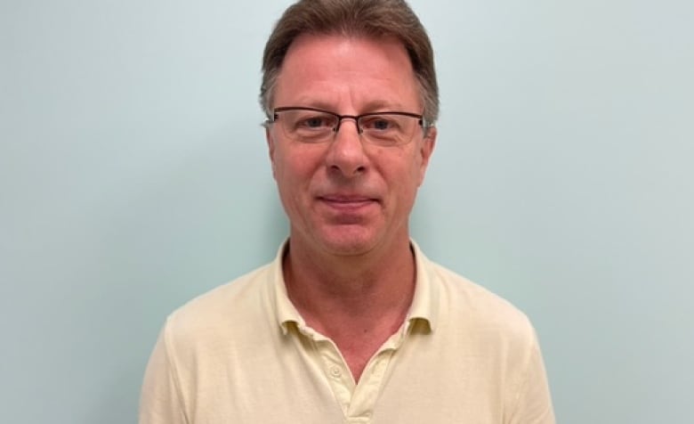 Allan Blyt stands with a blue wall behind him and looks at the camera. 