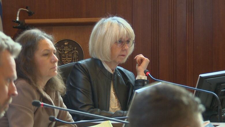 A woman with blond hair and wearing a black jacket sits behind a council desk in front of a microphone.