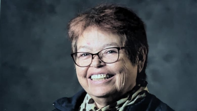 A smiling woman in her late 50s, with shorter reddish-brown hair, dark-rimmed glasses, wearing a darker jacket and a green scarf with floral print.