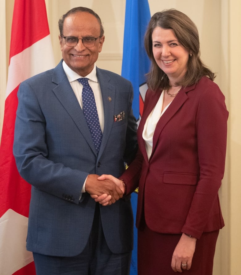 a man and a woman stand in front of two flags. The flags are of canada and alberta. They are smiling and shaking hands.