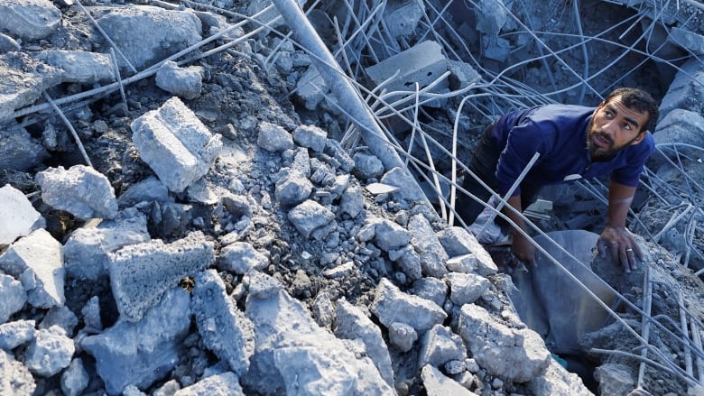 A bearded man in a tshirt is shown crouching, surrounded by rocks and concrete debris.