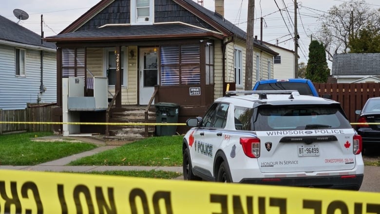 A Windsor police cruiser on the other side of yellow caution tape and parked in front of a one and a half storey house with the number 2415 on it and a beware of dog sign