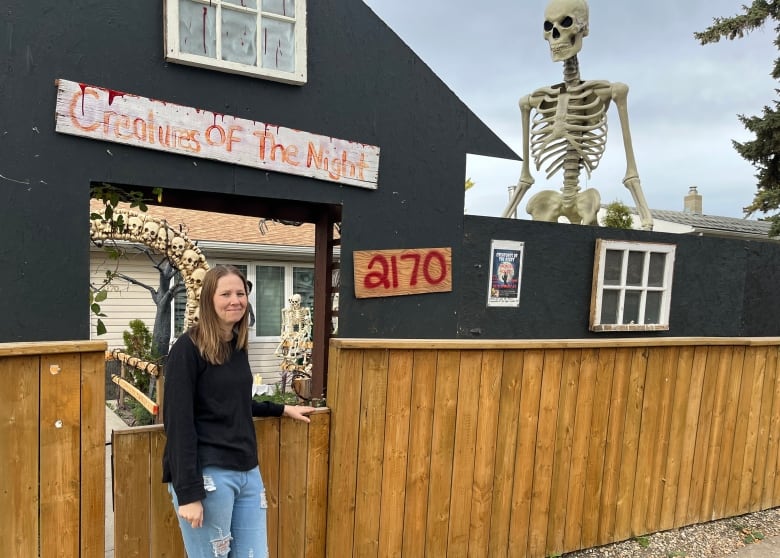 A woman wearing a black shirt and jeans stands in front of a wooden structure that looks like a house. Used to block the view into her haunted house.