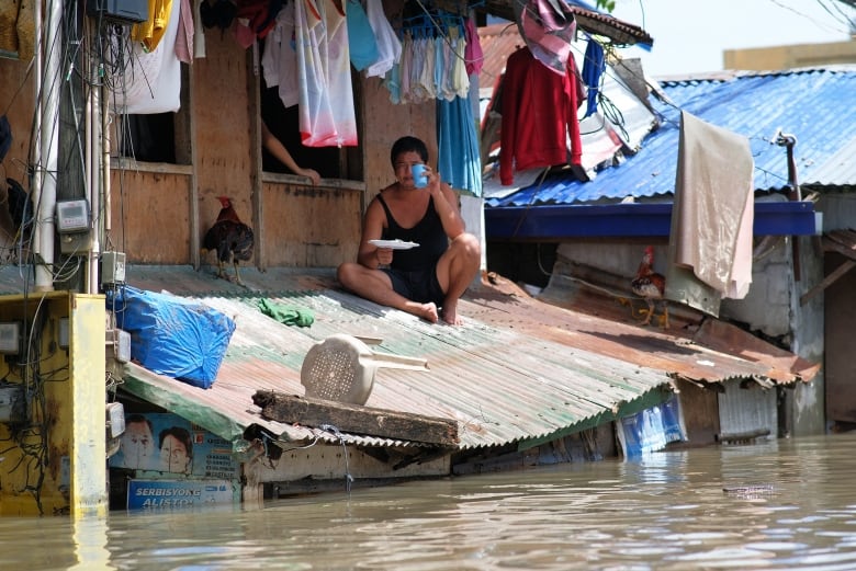 A person sits on a corrugated roof and eats as murky water has risen just below the roof level. Above the person, clothes hang out on a line.