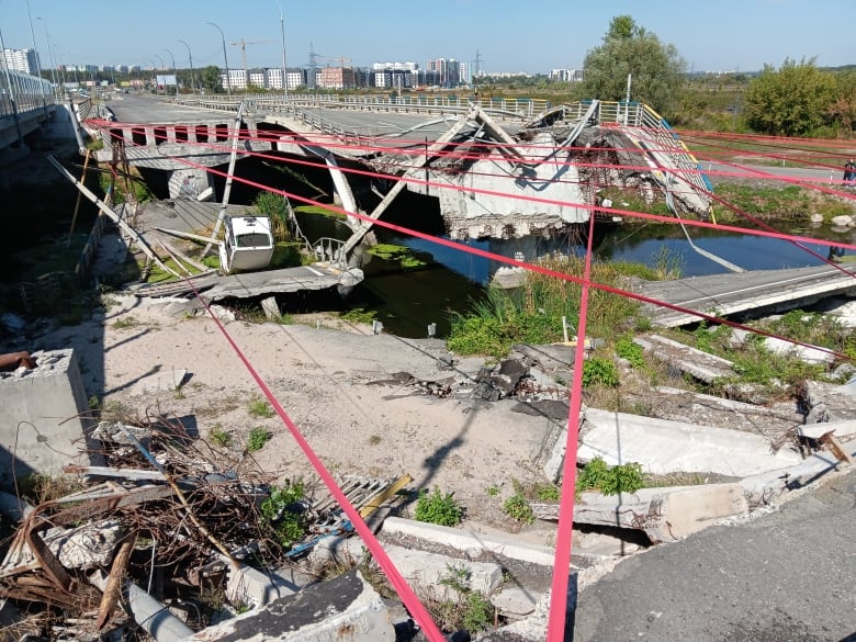 The ruins of what appears to be a bridge. A bus can be seen lying upside down in the wreckage. Red tape expands across the area. 