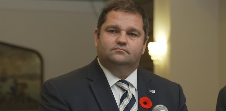 Close up of man in dark blue suit with striped blue tie. He's staring off centre. On his lapel is a red poppy pin and pin of Newfoundland and Labrador.