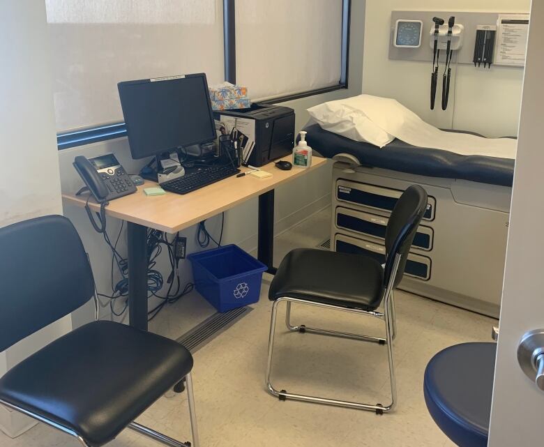 An empty medical examination room includes a patient examination bed, a desk with computer and two chairs. Medical equipment is hanging on the wall.