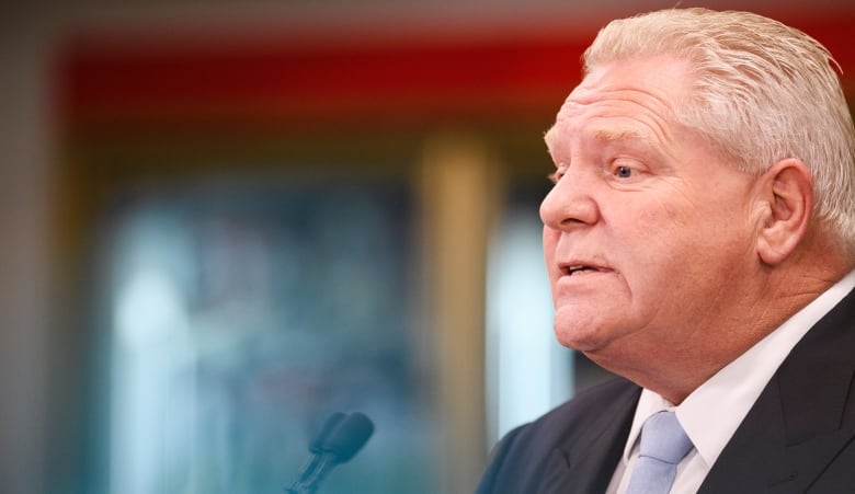 Close-up of a man's face in the middle of talking, shown in profile view in front of a blurred background. 