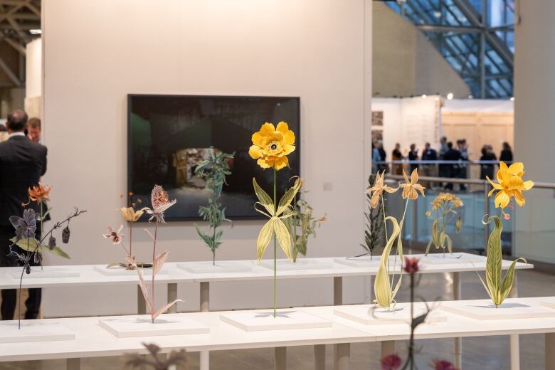 Photo of paper flowers standing on white tables in a crowded room.