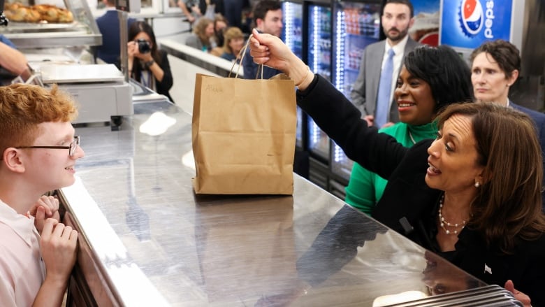 A dark-haired woman, wearing a black jacket, reaches up to get a paper bag off a counter, while a young man with red hair and glasses smiles.