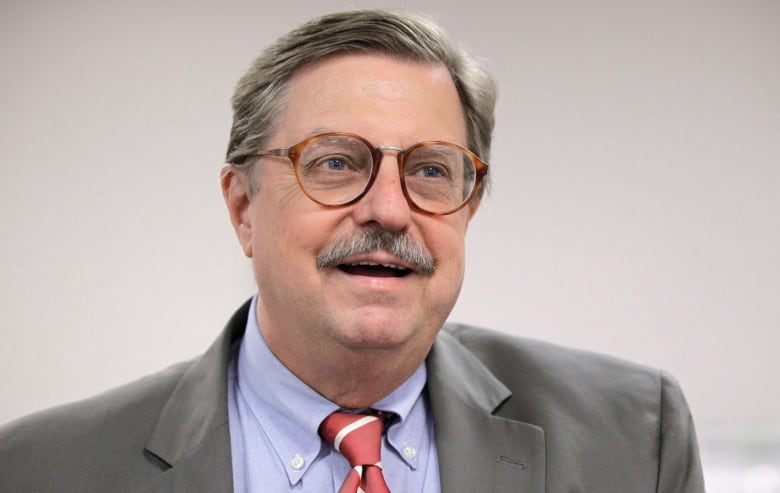 A man with grey hair and a moustache wears glasses, a grey jacket, light blue shirt and a red and white striped tie.