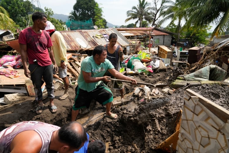 A man stands on top of a pile ruble from a landslide and uses a shovel while others watch.