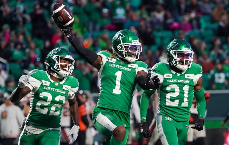 A football player holds up the ball in a victorious way on the field with two teammates.