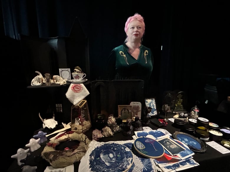 A woman dressed in a pink wig and a a dark green dress stands in front of a table filled with spooky-themed products. 