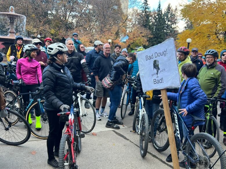 A few dozen cyclists gather outside in a park on a fall day. One cyclist near the front holds a protest sign.