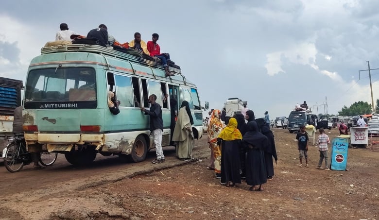 People wait to board a vehicle as others sit inside and atop it.