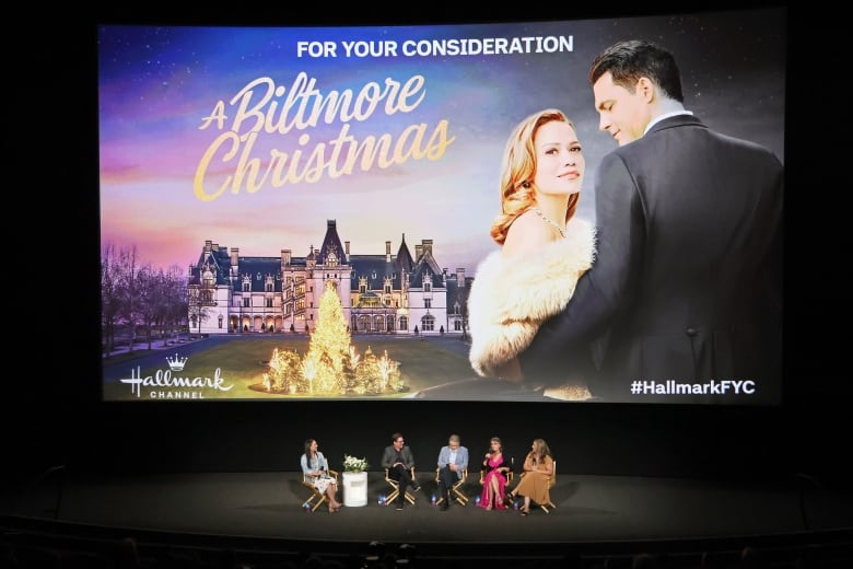 Five people sit on a stage, behind them is a giant projection for a  movie called A Biltmore Christmas