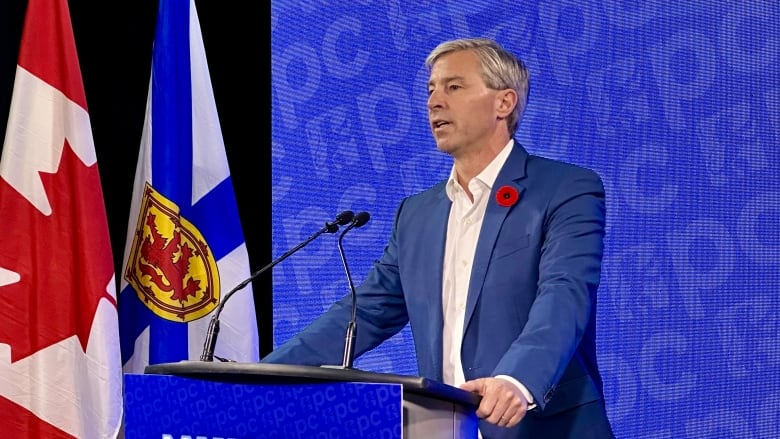 A man speaks at a podium in front of a blue screen. Canada and Nova Scotia flags hang beside him.